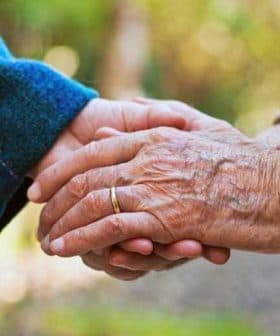 Two elderly hands clasped together, one wearing a ring and the other with a textured sweater sleeve. - Olive Oil Times