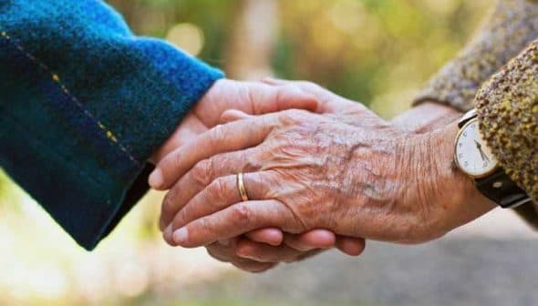 Two elderly hands clasped together, one wearing a ring and the other with a textured sweater sleeve. - Olive Oil Times