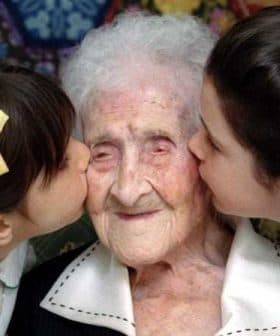 An elderly woman is being kissed on the cheeks by two young girls, one on each side, both smiling. - Olive Oil Times