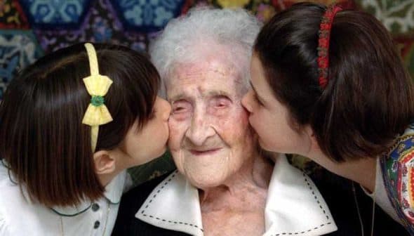 An elderly woman is being kissed on the cheeks by two young girls, one on each side, both smiling. - Olive Oil Times