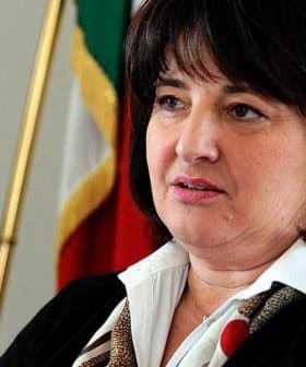 Woman with dark hair and a white shirt speaking in front of flags. - Olive Oil Times