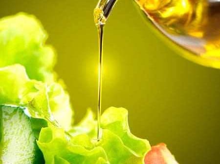 Olive oil being poured from a glass container onto a fresh green salad with lettuce and vegetables. - Olive Oil Times