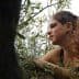 A woman with curly hair sitting among olive tree branches, looking thoughtfully to the side. - Olive Oil Times