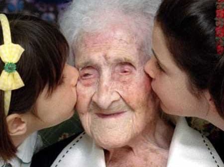 An elderly woman is being kissed on the cheeks by two young girls on either side of her. - Olive Oil Times
