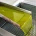 Stream of green olive oil flowing from a stainless steel spout into a perforated container. - Olive Oil Times