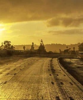 A wet road with reflective surfaces during sunset, featuring distant trees and signage. - Olive Oil Times