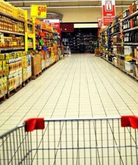 View of a supermarket aisle with shelves stocked with various products and a shopping cart in the foreground. - Olive Oil Times