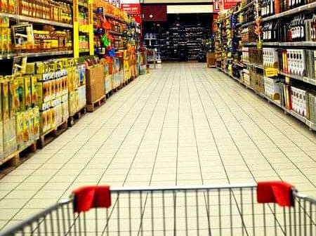 View of a supermarket aisle with shelves stocked with various products and a shopping cart in the foreground. - Olive Oil Times