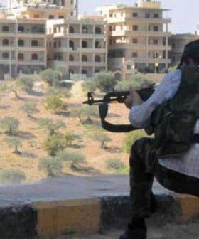 Individual in military attire aiming a firearm while crouched down in a rural area with buildings in the background. - Olive Oil Times