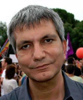 Man with short gray hair and a neutral expression at a public gathering with people in the background. - Olive Oil Times