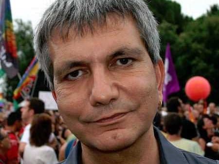 Man with short gray hair and a neutral expression at a public gathering with people in the background. - Olive Oil Times