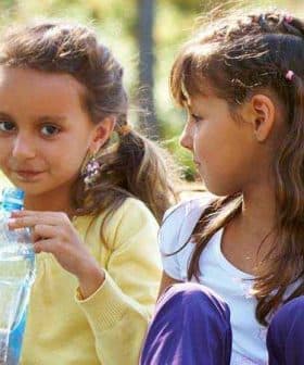 Two young girls sitting outdoors, one holding a plastic water bottle and looking at the other. - Olive Oil Times