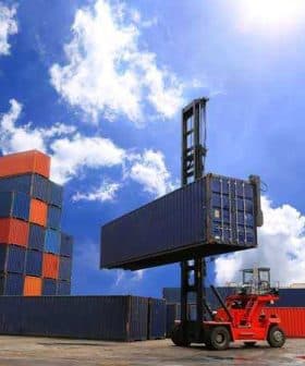 Red forklift lifting a blue shipping container at a cargo yard with stacked containers in the background. - Olive Oil Times