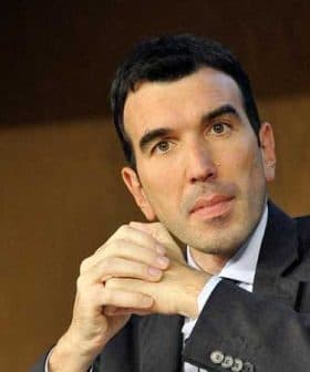 A man in a dark suit with short black hair, sitting with his hands clasped in front of him. - Olive Oil Times