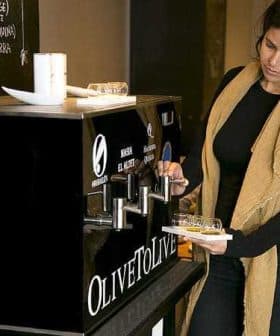 A woman dispensing olive oil from a black dispenser labeled 'OLIVETO LIVE' in a casual setting. - Olive Oil Times