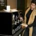 A woman dispensing olive oil from a black dispenser labeled 'OLIVETO LIVE' in a casual setting. - Olive Oil Times