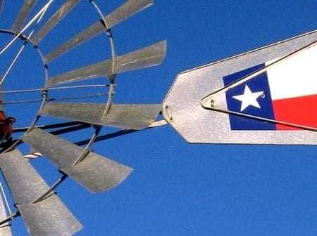 Close-up of a windmill with a Texas state flag attached to its arm against a blue sky. - Olive Oil Times