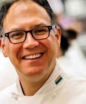 Chef wearing a white uniform and glasses, smiling at the camera in a kitchen setting. - Olive Oil Times