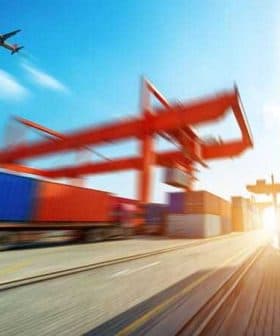 Cargo containers stacked at a shipping port with an airplane flying overhead in a clear sky. - Olive Oil Times