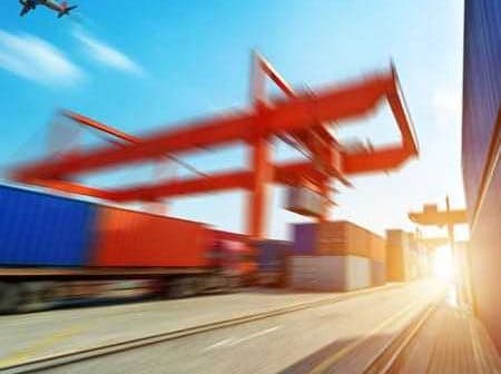 Cargo containers stacked at a shipping port with an airplane flying overhead in a clear sky. - Olive Oil Times