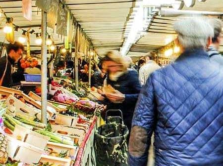Busy market scene featuring various stalls with fresh produce and shoppers walking through the aisles. - Olive Oil Times