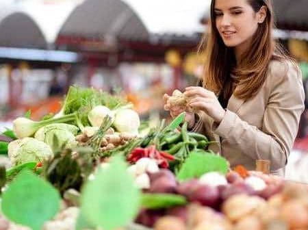 A woman examining fresh vegetables at a market stall filled with various produce. - Olive Oil Times