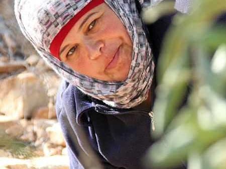 Woman wearing a headscarf and a dark jacket, smiling while harvesting olives in a field. - Olive Oil Times