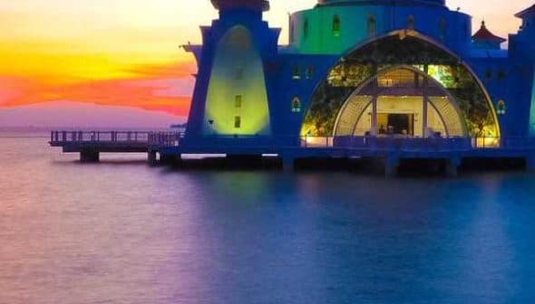 A mosque with a golden dome and colorful lighting situated by the water during sunset. - Olive Oil Times