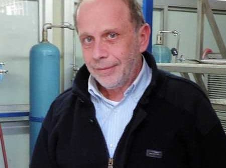 A man wearing a black jacket and a light blue shirt, standing in an industrial setting. - Olive Oil Times