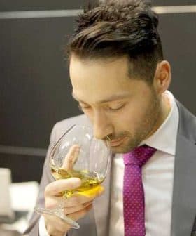 Man in a suit holding a glass of olive oil and smelling it at an event. - Olive Oil Times