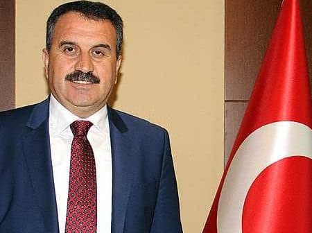 A man in a suit standing next to a Turkish flag, with a neutral expression and a mustache. - Olive Oil Times