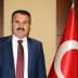 A man in a suit standing next to a Turkish flag, with a neutral expression and a mustache. - Olive Oil Times