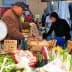 A market vendor handling fresh vegetables and fruits at a market stall with a customer selecting items. - Olive Oil Times