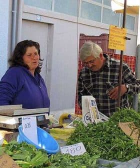 A market stall featuring various fresh herbs and vegetables with two individuals interacting. - Olive Oil Times