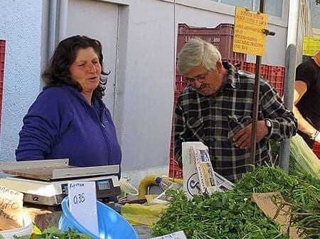 A market stall featuring various fresh herbs and vegetables with two individuals interacting. - Olive Oil Times