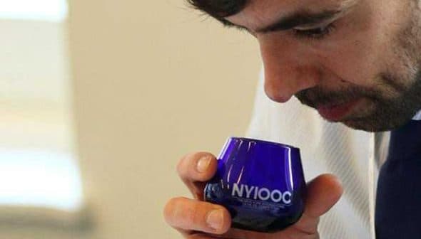 A man holding a blue glass with 'NYIOOC' while smelling its contents during an olive oil tasting. - Olive Oil Times
