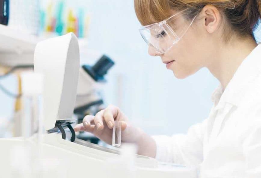 Female scientist in a lab coat and safety glasses operating laboratory equipment with test tubes. - Olive Oil Times