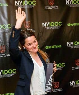 Woman with long hair raising her hand while holding an award at a culinary event backdrop. - Olive Oil Times
