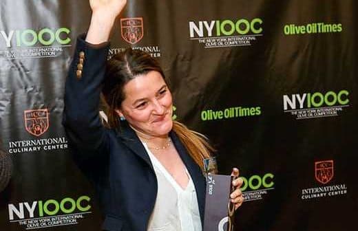 Woman with long hair raising her hand while holding an award at a culinary event backdrop. - Olive Oil Times