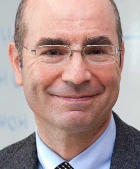 A man wearing glasses and a gray suit, smiling at the camera with a neutral background. - Olive Oil Times