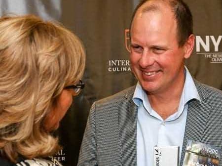 Man in a checkered blazer conversing with a woman at a formal event. - Olive Oil Times