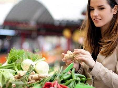 Woman examining fresh vegetables at a market stall with various produce visible. - Olive Oil Times