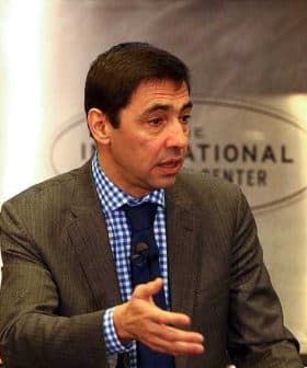 Man in a gray suit with a blue checkered shirt speaking at a podium during an event. - Olive Oil Times