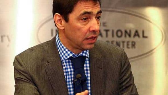 Man in a gray suit with a blue checkered shirt speaking at a podium during an event. - Olive Oil Times