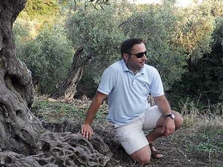 A man wearing sunglasses and a light shirt kneeling beside an olive tree in a natural setting. - Olive Oil Times