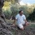 A man wearing sunglasses and a light shirt kneeling beside an olive tree in a natural setting. - Olive Oil Times
