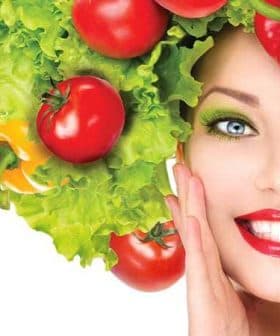 Woman with a hairstyle made of lettuce and tomatoes, smiling and posing with a hand on her face. - Olive Oil Times