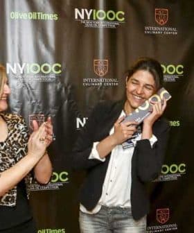 A woman smiling while holding an award at an event with a backdrop featuring Olive Oil Times and NYIOOC logos. - Olive Oil Times