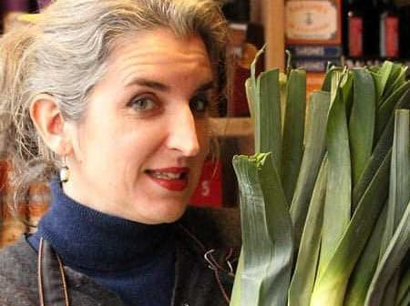 Woman with gray hair holding a bunch of fresh leeks in a market setting. - Olive Oil Times