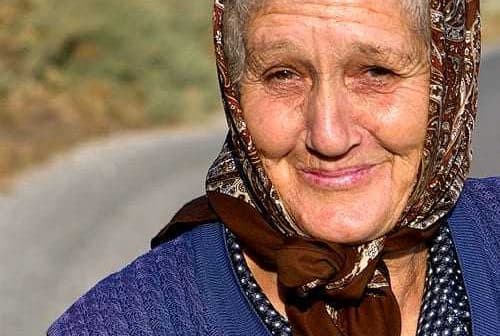 Elderly woman wearing a patterned headscarf and a blue vest, smiling at the camera. - Olive Oil Times